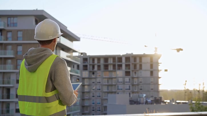 acilities manager with a white hard hat and safety vest is using a tablet computer while inspecting a building at sunset.